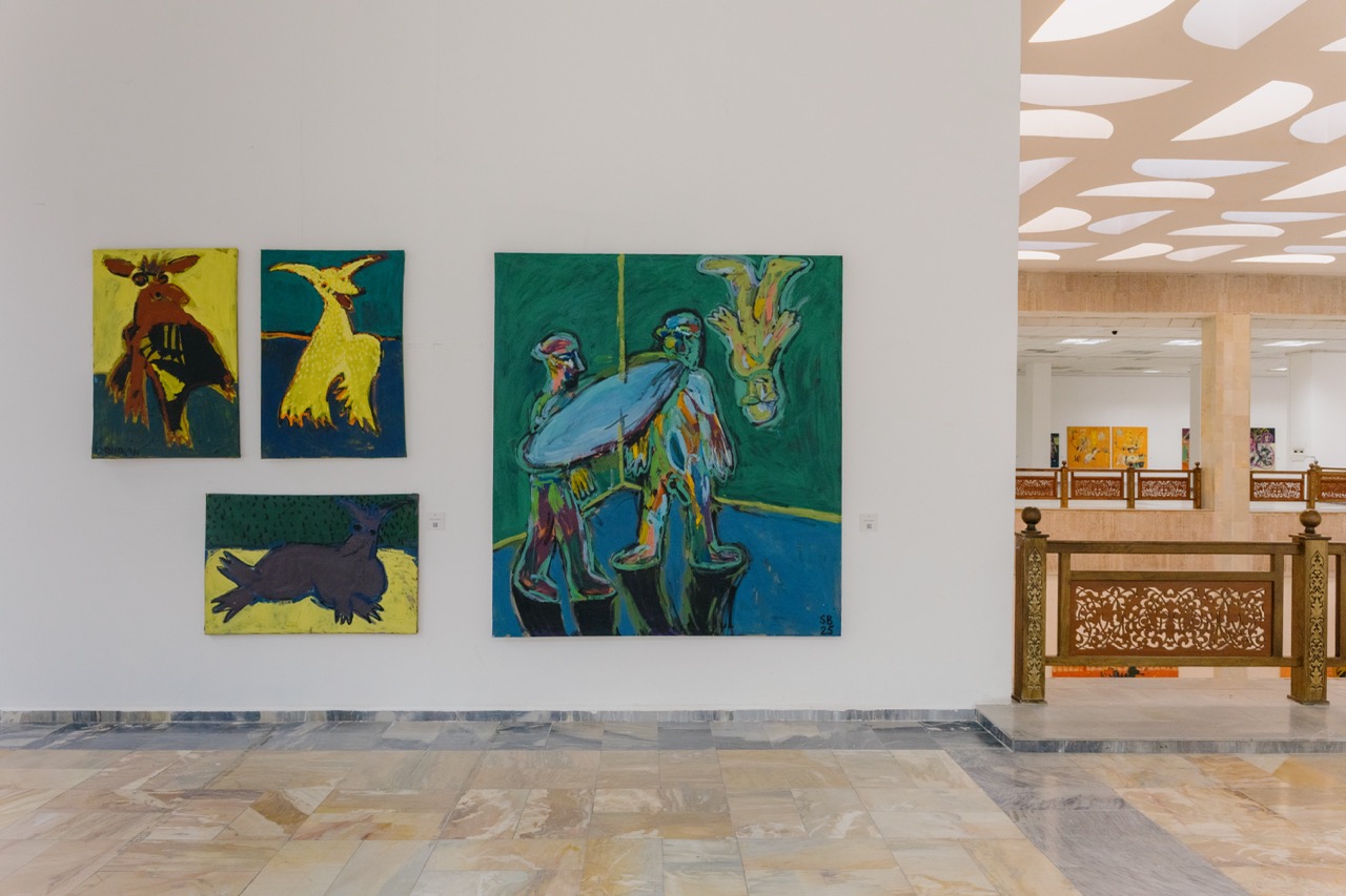 Photo of well-lit bright exhibition hall with tiled floor, with paintings hung up on walls