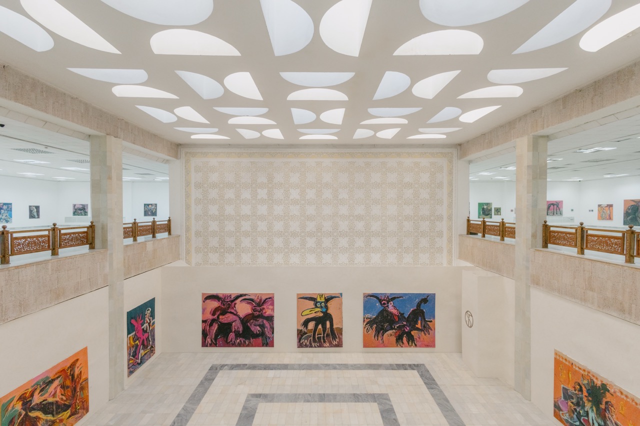Photo of well-lit bright exhibition hall with tiled floor, with paintings hung up on walls