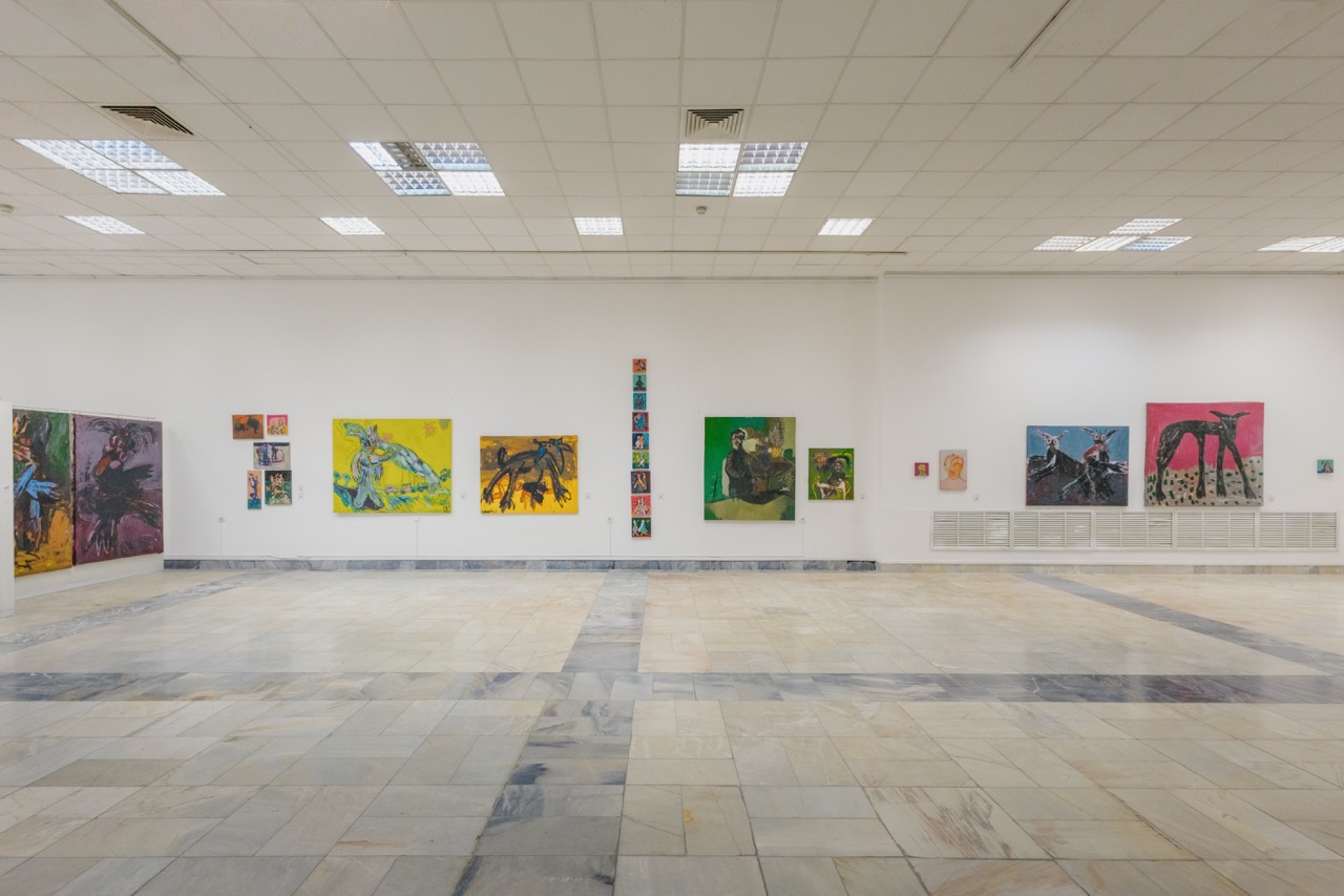Photo of well-lit bright exhibition hall with tiled floor, with paintings hung up on walls