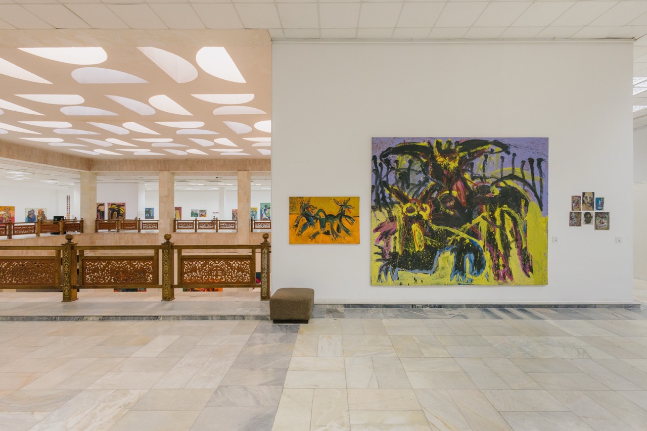 Photo of well-lit bright exhibition hall with tiled floor, with paintings hung up on walls